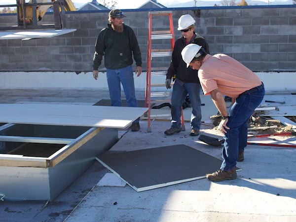 TPO-Roof-Installation-Canyon-Lake-CA-California-2 | Woolbright's Roofing & Construction Three construction workers on a flat roof wearing hard hats, bending to fit a white panel while others watch; a red ladder and skylight frame nearby.